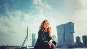 Vrouw in pak hurkt bij water, skyline en Erasmusbrug op achtergrond, bewolkte lucht.