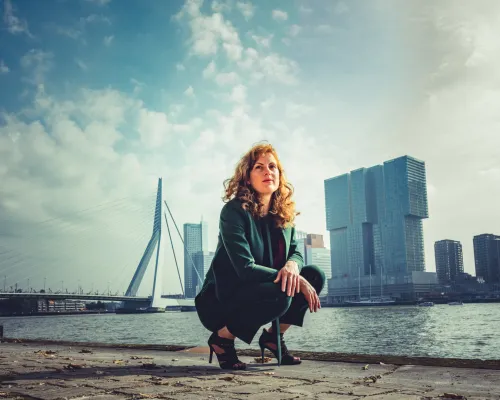 Vrouw in pak hurkt bij water, skyline en Erasmusbrug op achtergrond, bewolkte lucht.